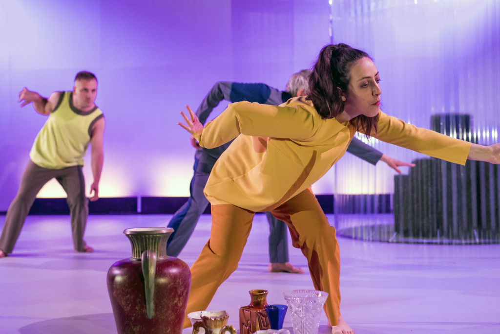 Szenenfoto aus einer Auffuehrung: Drei Menschen tanzen auf einer Theaterbühne. Im Vordergrund Blumenvasen.