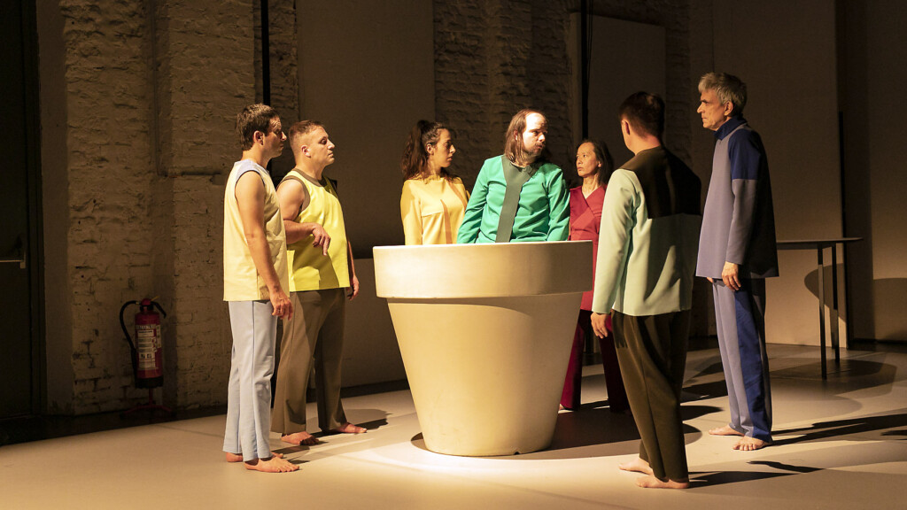 Scene photo of a performance: A couple people stand around a flower pot. one is in the flower pot.