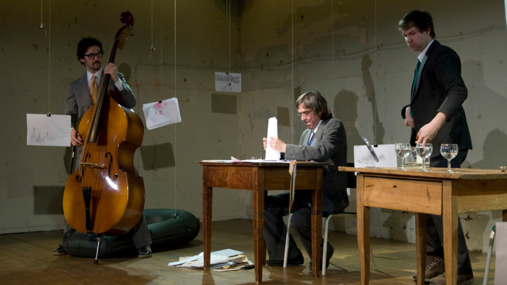Szenenfoto aus einer Auffuehrung: Links ein Mann mit Kontrabass. Rechts daneben sitzt und steht jeweils ein Mann an einem Holztisch.