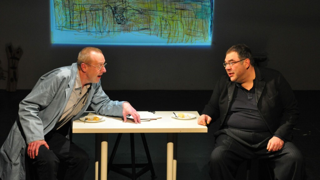 Photo from a performance: Two men converse on a table on a stage.