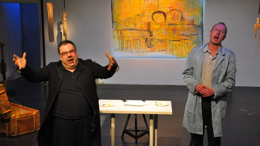 Szenenfoto aus einer Auffuehrung: Zwei Männer auf einer Theaterbühne. Im Hintergrund ein Tisch und eine Leinwand.