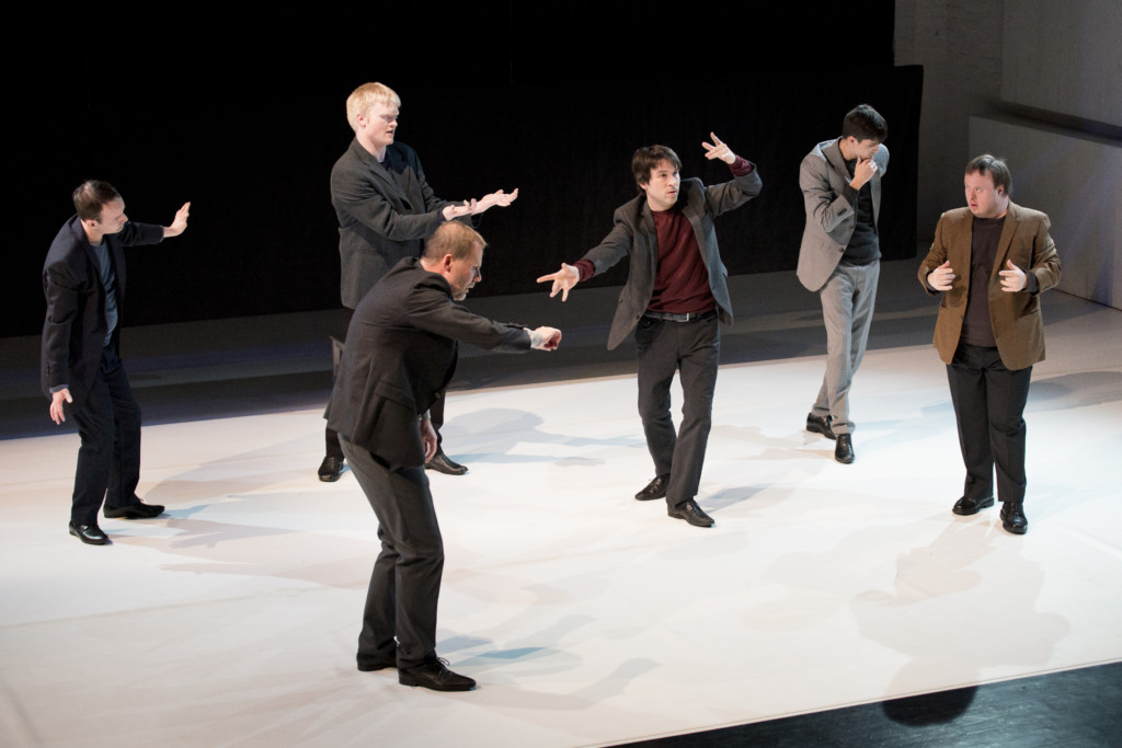 Szenenfoto aus einer Auffuehrung: Sechs Männer in Anzügen stehen auf einer Theaterbühne.