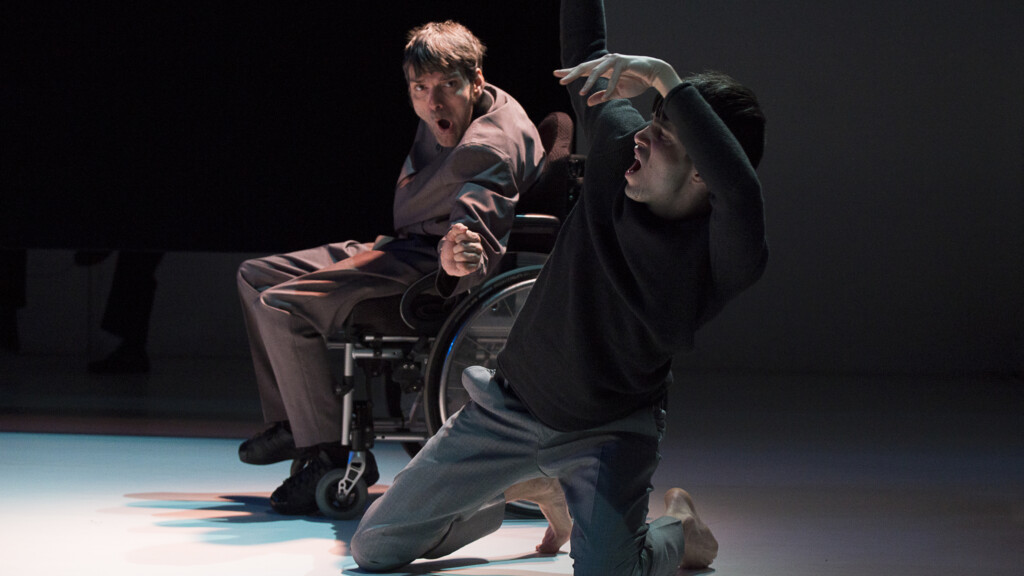 Scene photo of a performance: Two men on a stage. One kneeling in the foreground. The other one is sitting in a wheelchair in the background.