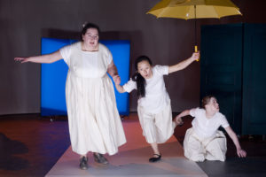 Szenenfoto aus einer Auffuehrung: Drei Frauen auf der Bühne. Zwei stehen und eine kniet.