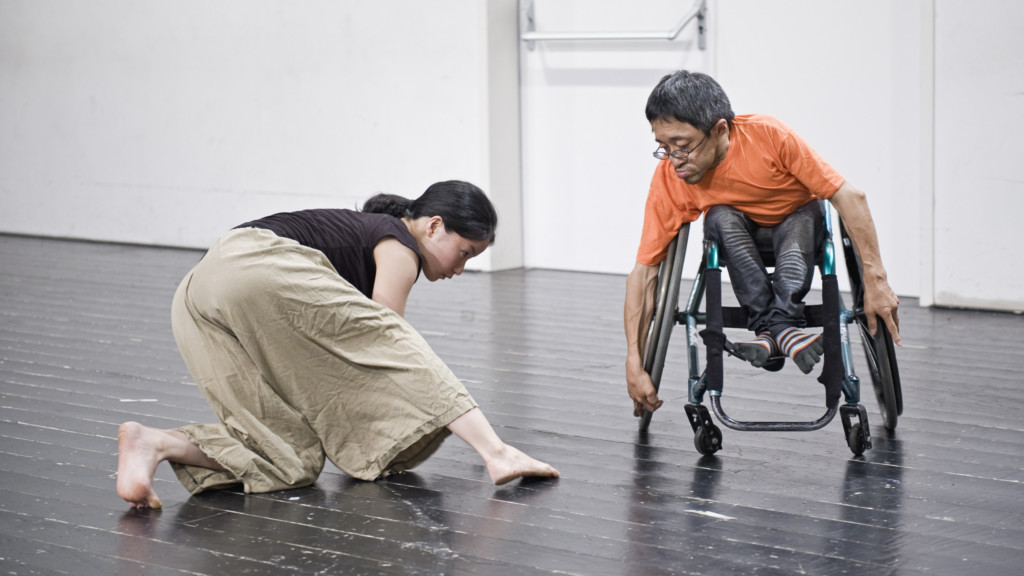 Szenenfoto: Ein Mann im Rollstuhl und eine Frau auf Knieen in Bewegung. Beide sind japanisch.