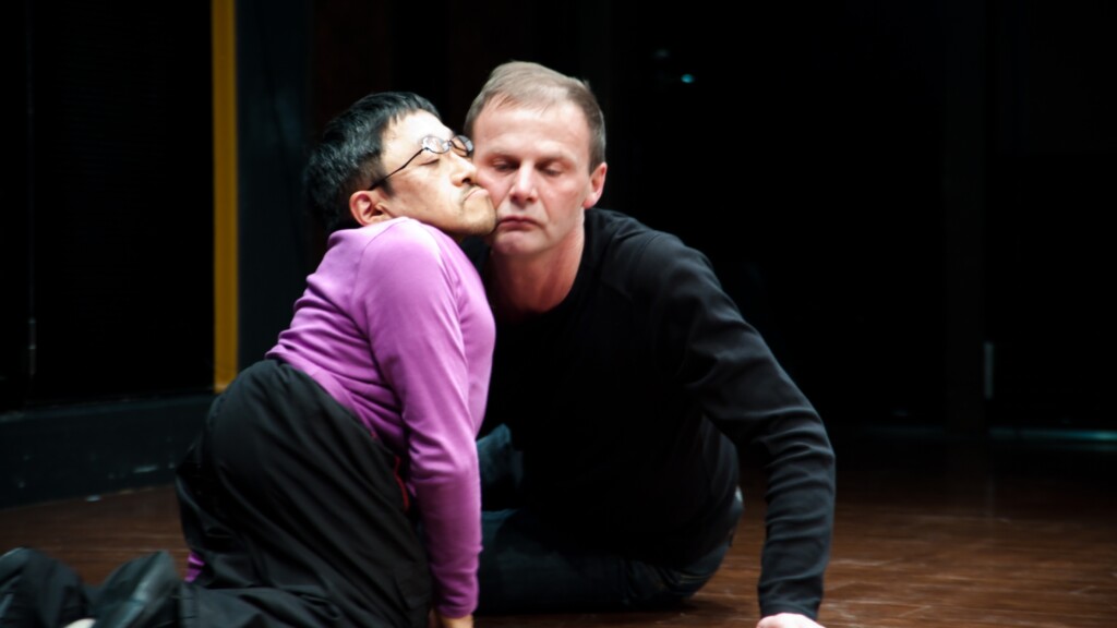 Scene photo of a performance: Two men on a stage. Their cheeks touching each other.