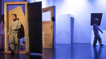 Szenenfoto aus einer Aufführung: Links eine offene Holztür, die ein Mann im Overall durchschreitet. Rechts im Hintergrund eine Frau im Overall mit einem rechteckigen Gebilde am Kopf.