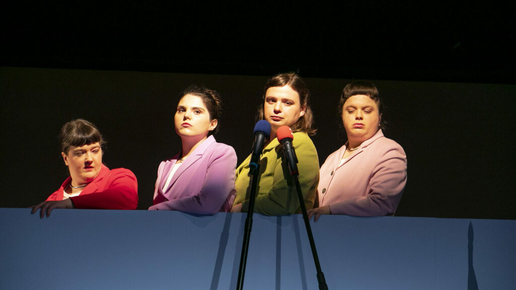 Scene photo of a performance: 4 women on a stage wearing blazers standing behind each other.