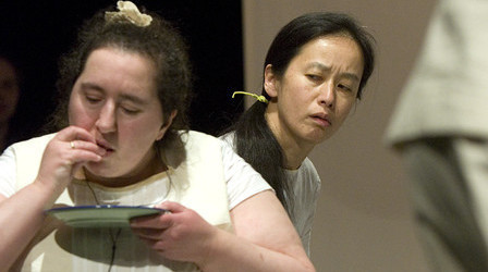 Szenenfoto aus einer Auffuehrung: Zwei Frauen. Eine isst etwas von einem Teller. Die andere beobachtet sie dabei