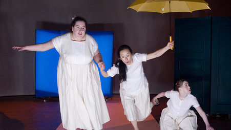 Szenenfoto aus einer Auffuehrung: Drei Frauen, weiß gekleidet. Eine hat einen gelben Regenschirm