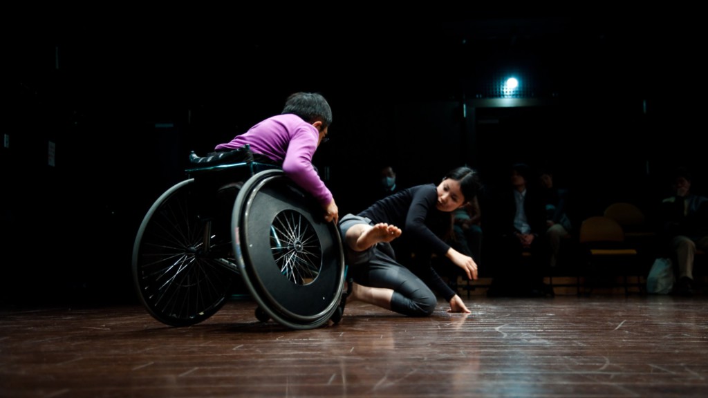 Szenenfoto aus einer Auffuehrung: Ein Mann im Rollstuhl und eine junge Frau tanzen akrobatisch