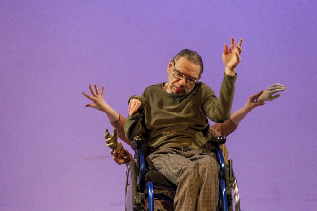 Scene photo of a performance: A man in a wheelchair on a stage. Other people are being him. But you just see their hands.