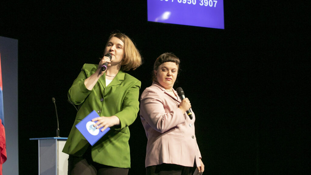 Scene photo of a performance: Two performers with microphones. In the background a telephone number on a screen.