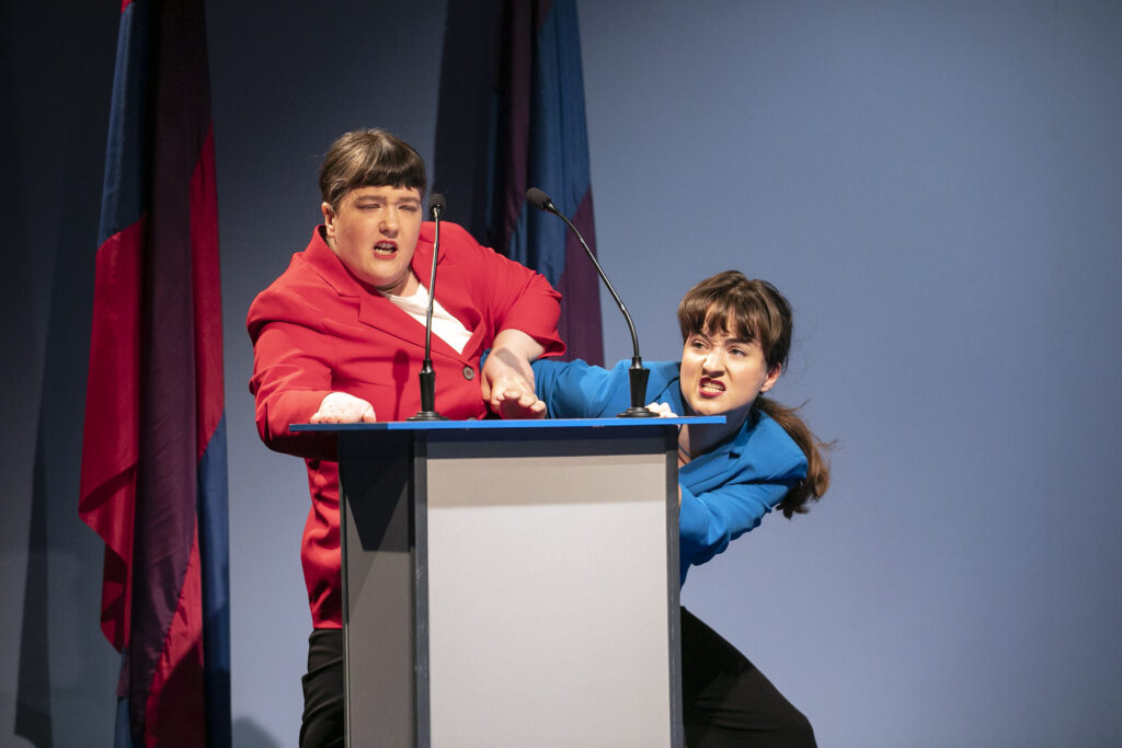 Szenenfoto einer Auffuehrung: Zwei Performerinnen streiten sich um einen Platz am Rednerpult.