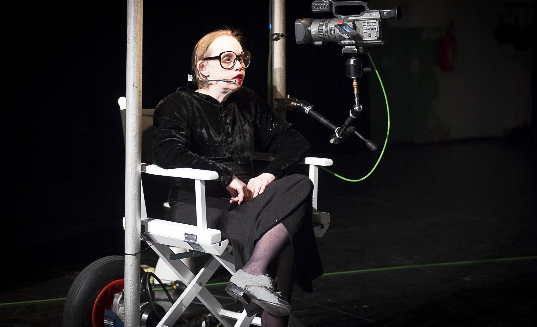 Scene photo of a performance: One woman is sitting in a directors chair.