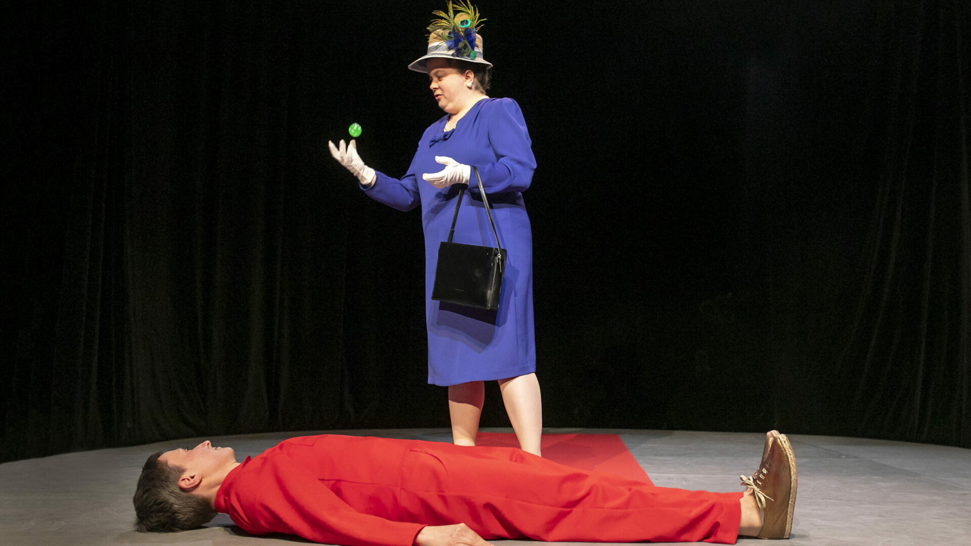 Scene photo of a performance: One person is lying on her back on the ground. She is looking at the other person behind her who throws a bouncy ball in the air with her right hand.