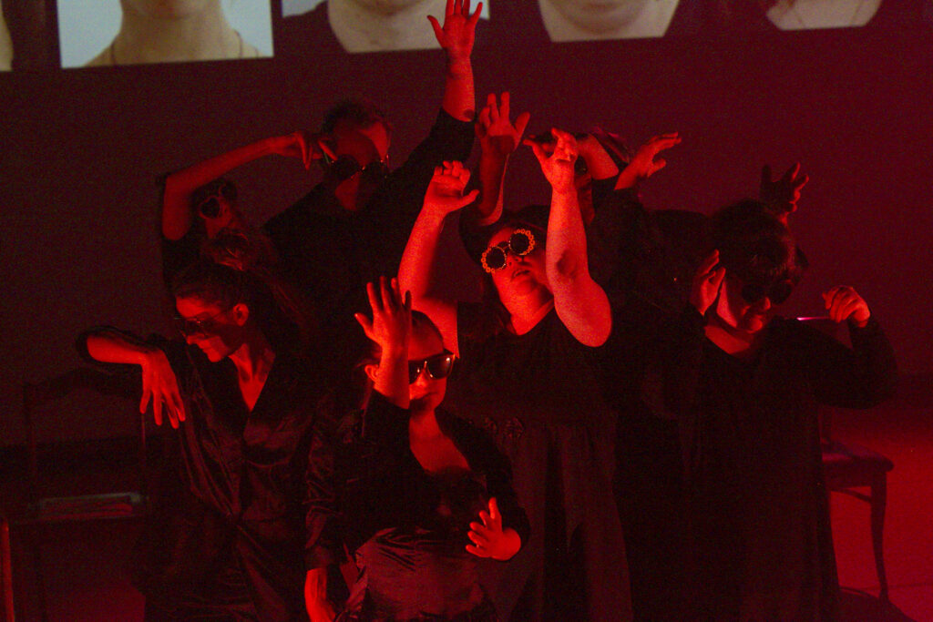 Szenenfoto einer Auffuehrung: Mehrere Personen tanzen dicht beieinander auf einer Buehne. Sie heben ihre Arme.
