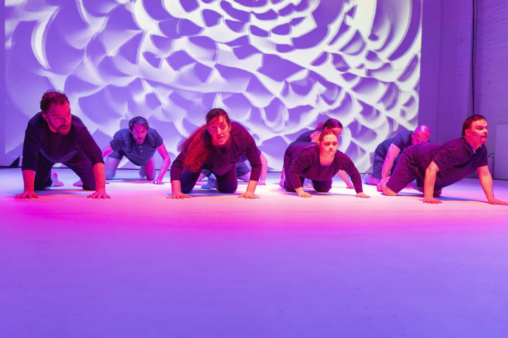 Eine lila angeleuchtete Bühne auf der sechs Personen auf dem Boden kauern.