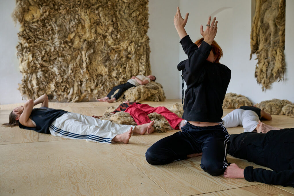 Ein Raum mit hölzernem Boden und roher Schafwolle an der Wand und auf dem Boden. Mehrere Personen liegen auf dem Boden, eine kniet und hält die Arme verschränkt vor ihrem Gesicht.