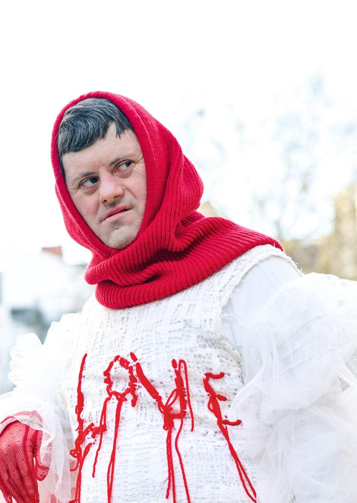 Ein Mann mit einer roten Kapuze und einem weißen Gewand steht vor einer Absperrung. Auf seinem weißen Oberteil ist "Love" gestickt.